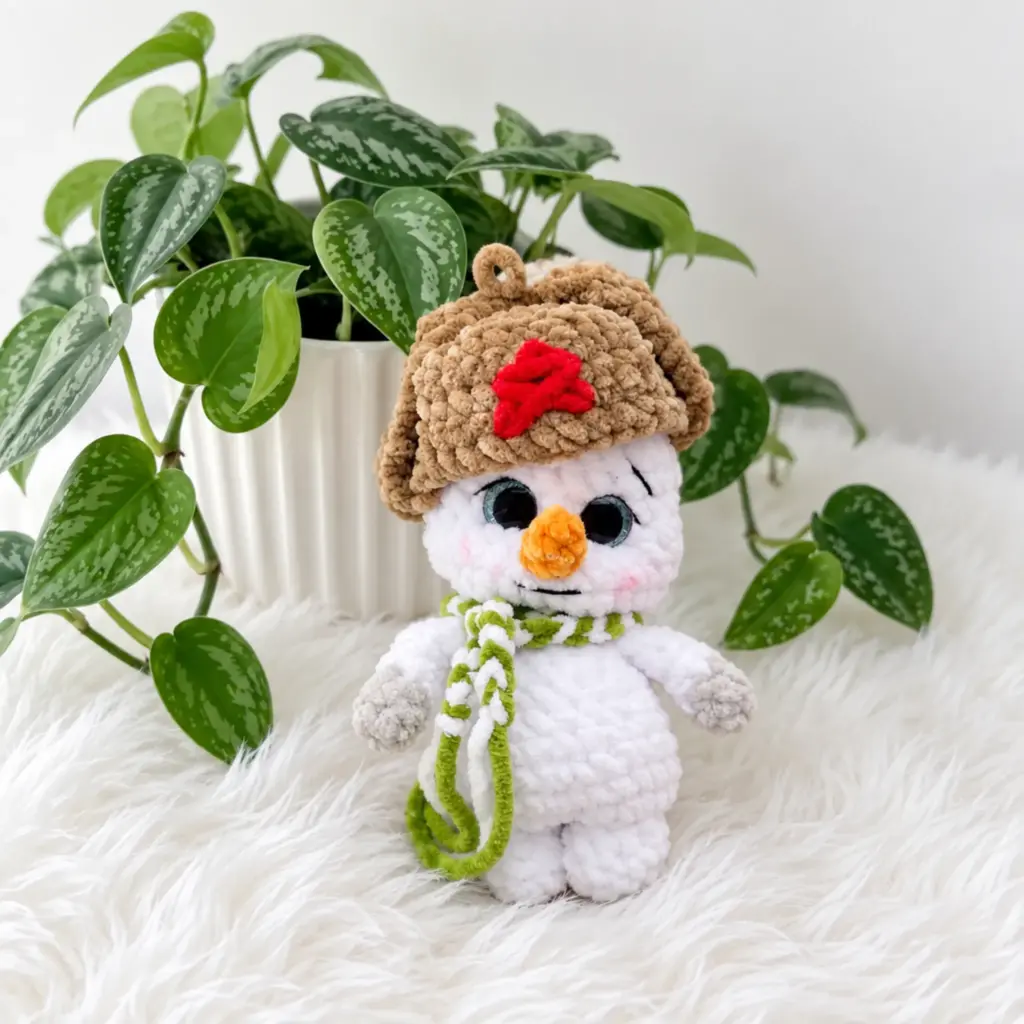 Finished ushanka snowman amigurumi wearing its crochet hat and scarf, sitting beside a potted plant